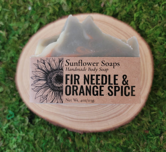 A handmade bar of Sunflower Soaps body soap labeled 'FIR NEEDLE & ORANGE SPICE' resting on a round wooden slice over a green grass background.