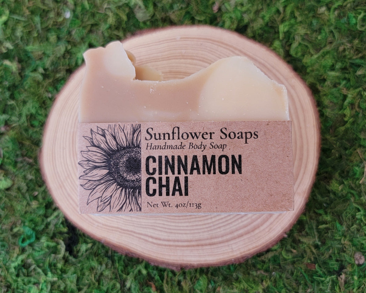 A bar of Sunflower Soaps Cinnamon Chai handmade body soap rests on a round wooden slice, set against a background of green moss. The soap bar has a natural cream-colored texture and is wrapped in a kraft paper label featuring a black sunflower illustration.