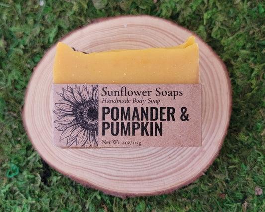 A bar of Sunflower Soaps Pomander & Pumpkin handmade body soap, wrapped in kraft paper, resting on a round wooden slice over a green moss background.